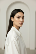 Woman in white outfit standing against neutral beige background wearing delicate champagne gold four-leaf clover stud earrings with white mother-of-pearl inlay, minimal jewellery styling with soft natural lighting and warm undertones