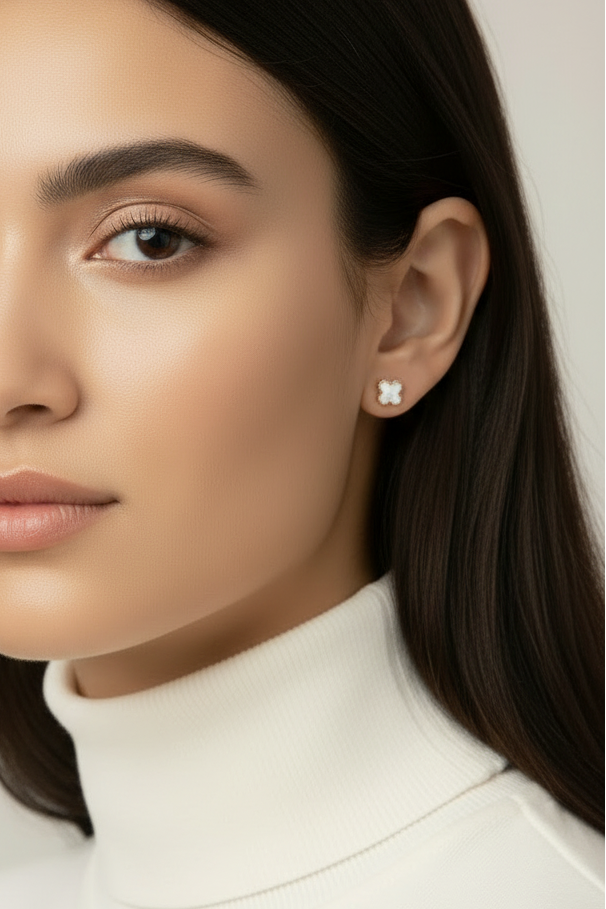 Close-up profile view of woman wearing champagne gold clover stud earrings with white mother-of-pearl inlay against neutral beige background, showing earring positioned on earlobe with natural skin tones, soft lighting highlighting the delicate four-petal clover design and polished gold finish