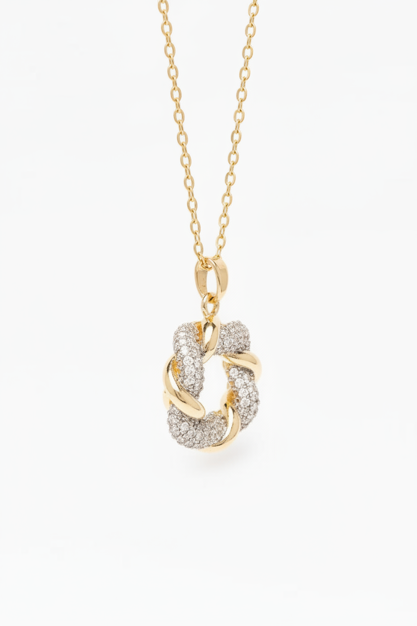 Detail view of diamond knot pendant in champagne gold with pavé crystal detailing on thin chain against cream background, refined minimal craftsmanship showing intricate knot construction and polished metallic finish