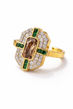 Gold ring with a central brown gemstone, surrounded by smaller diamonds and green gemstones on a white background
