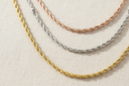 Tri-tone twisted chain necklace set in rose gold, silver and champagne gold arranged on textured cream linen fabric, flat lay composition showing three separate chains of varying lengths, minimal elegant design with warm beige aesthetic and polished metallic finish