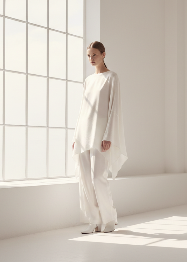Person wearing a white outfit standing in a bright room with large windows.